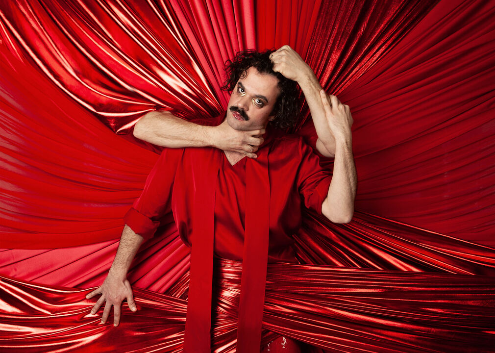 Een man in een rode outfit poseert te midden van een achtergrond van rode stoffen, met een hand om zijn nek en een andere hand op zijn hoofd.